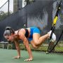 17378PS_01
A fit woman performing a suspended core exercise using TRX straps outdoors. Her feet are secured in the straps while she supports her body weight on her hands, engaging her core. The background features a chain-link fence and a green outdoor w thumbnail