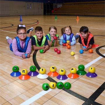 Small colored balls in the foreground; resting on mini cones on a gym floor; spelling out the words brainball mini. A group of diverse elementary age children; laying on the floor behind the balls; each holding foam balls.