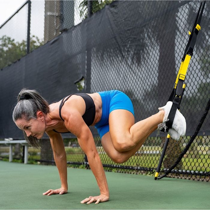 17378PS_01
A fit woman performing a suspended core exercise using TRX straps outdoors. Her feet are secured in the straps while she supports her body weight on her hands, engaging her core. The background features a chain-link fence and a green outdoor w
